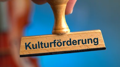 Ein symbolischer Holzstempel, gehalten von einer Hand mit der Aufschrift "Kulturförderung", mit einer Geige (unscharf) am Bildrand im Anschnitt; © picture alliance/SULUPRESS.DE/Torsten Sukrow